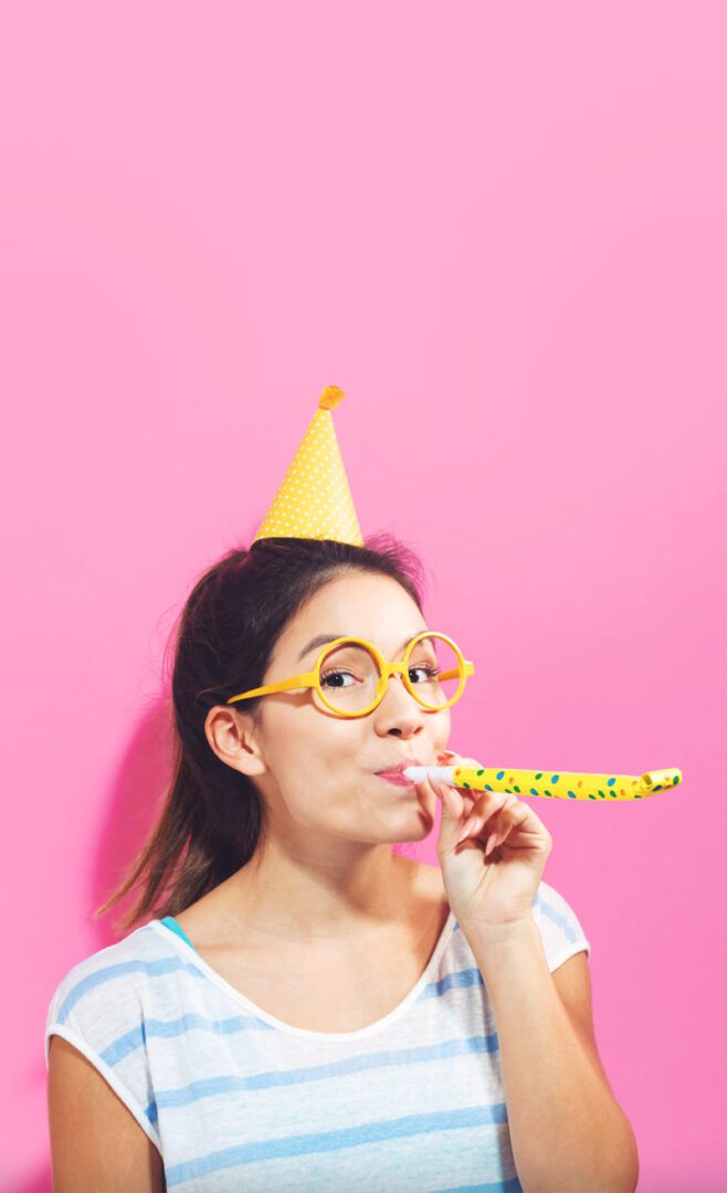 A woman wearing glasses and a party hat blowing on a straw.