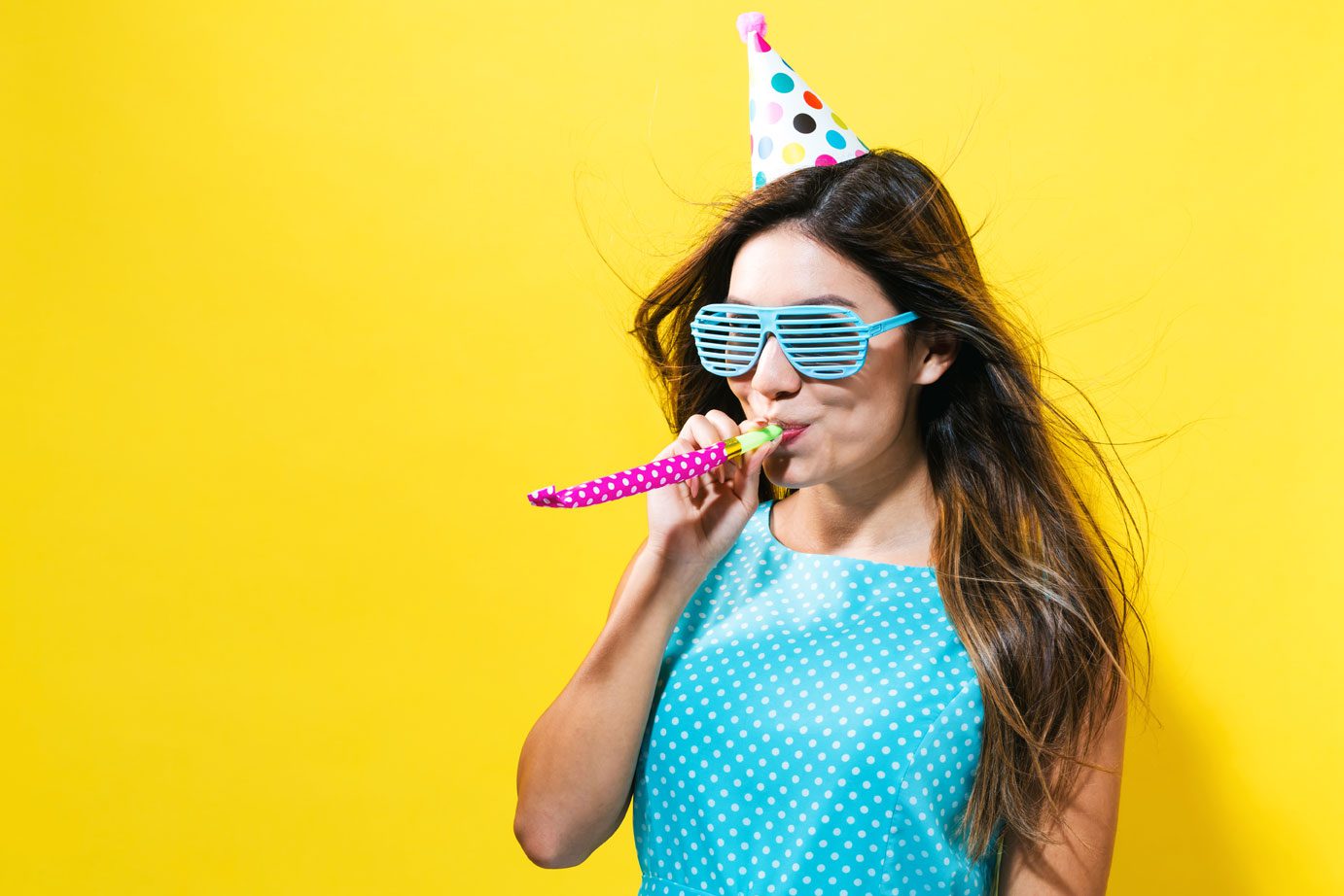 A woman wearing sunglasses and a party hat blowing on a pink horn.