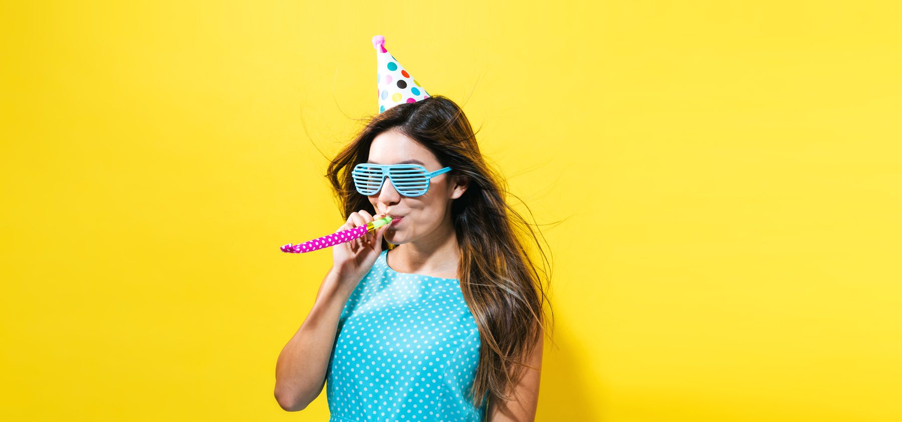 A woman wearing sunglasses and a party hat blowing on a blowfish.