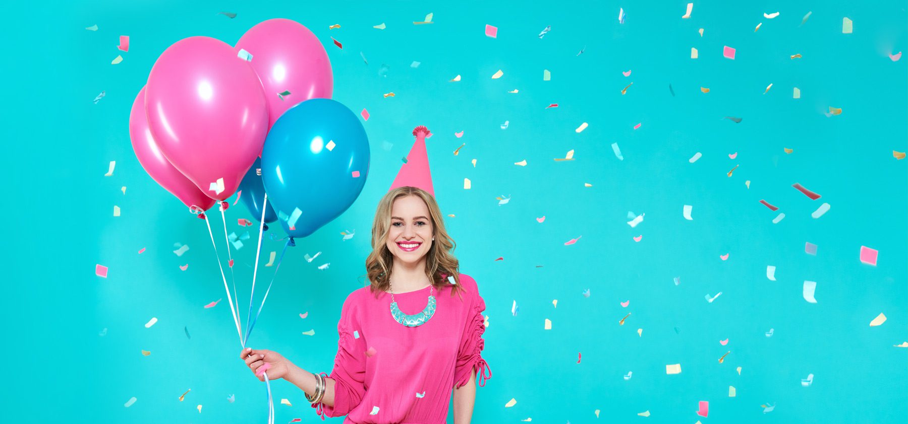 A woman in pink shirt holding balloons and wearing a party hat.