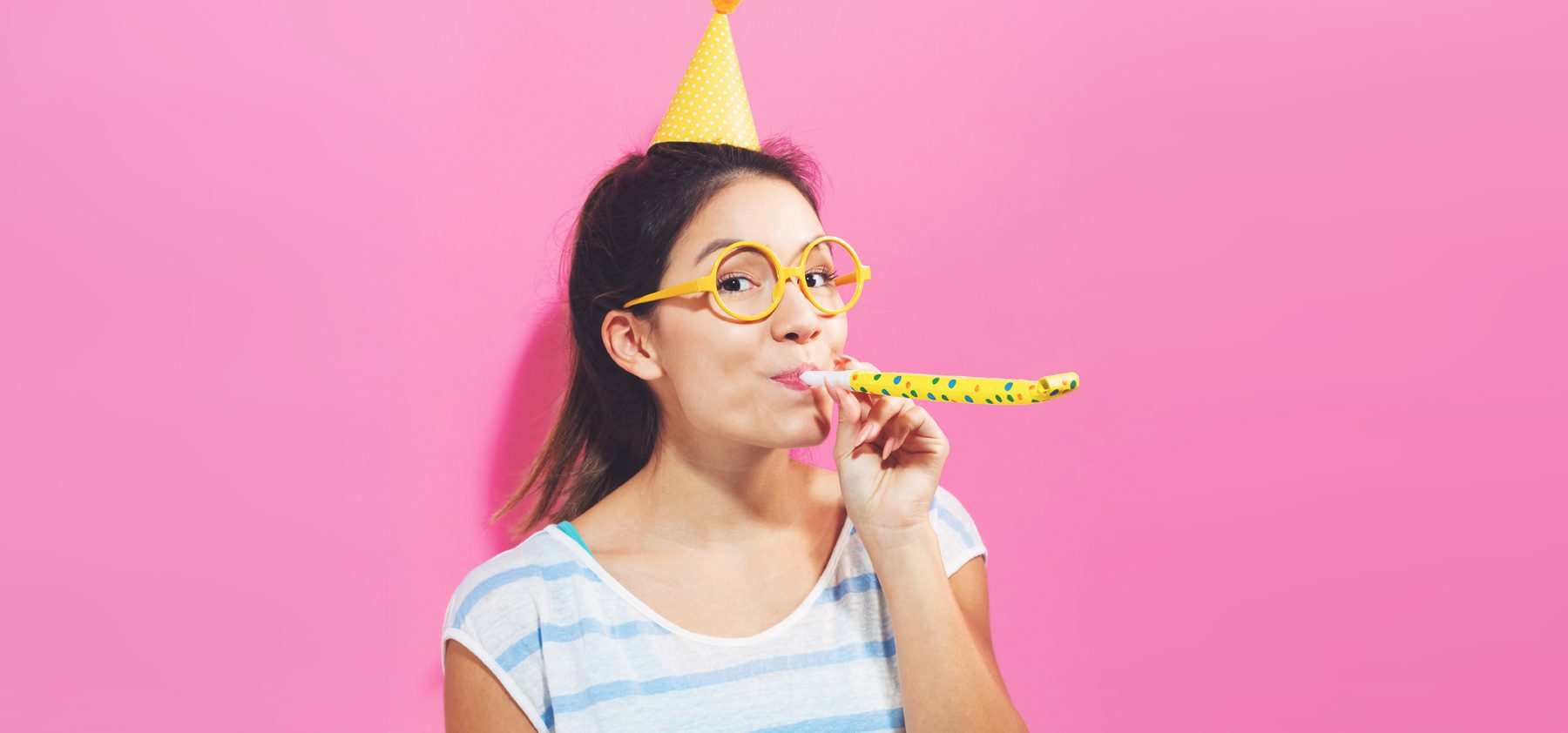 A woman wearing glasses and a party hat is holding a banana.