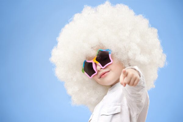 A child with white hair and sunglasses pointing at the camera.